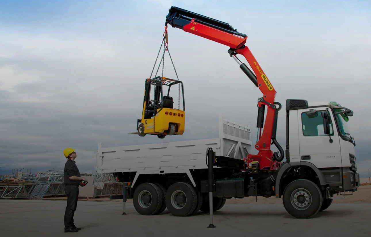 Un camion-grue déploie sa grue auxiliaire sur un chantier, son chauffeur au sol